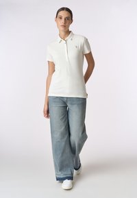 Polo bianco a maniche corte, con chiusura a bottoni e piccolo logo. Abbinato a jeans a gamba larga blu chiaro e sneakers bianche.