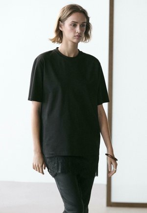 Young woman with short hair wearing a loose black t-shirt layered over a black lace-trimmed top and black pants, standing indoors.