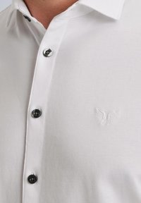 Weißes Hemd aus glattem Stoff mit spitzem Kragen, schwarzen Knöpfen und einem geprägten Adler-Logo auf der linken Brust.