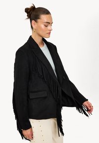 Femme avec les cheveux relevés en chignon, portant des boucles d'oreilles en or, un blazer noir à franges par-dessus une chemise grise, ainsi que des pantalons clairs avec des clous en or sur le côté.