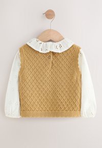 Gilet sans manches en maille beige pour enfant sur chemisier blanc à manches longues avec manches en dentelle à œillets et col à volants sur cintre en bois.