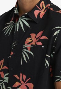 Chemise noire à manches courtes ornée de motifs floraux rouges et turquoise, col à boutons et une poche plaquée unique avec une étiquette logo.