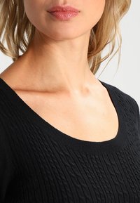 Ingrandimento del collo e delle labbra di una donna che indossa un maglione nero a coste con scollatura a barca su uno sfondo neutro.