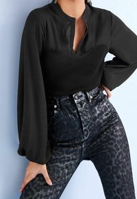 Blouse noire avec un col haut et un devant côtelé, associée à un jean noir ajusté arborant un imprimé léopard discret et une texture brillante.