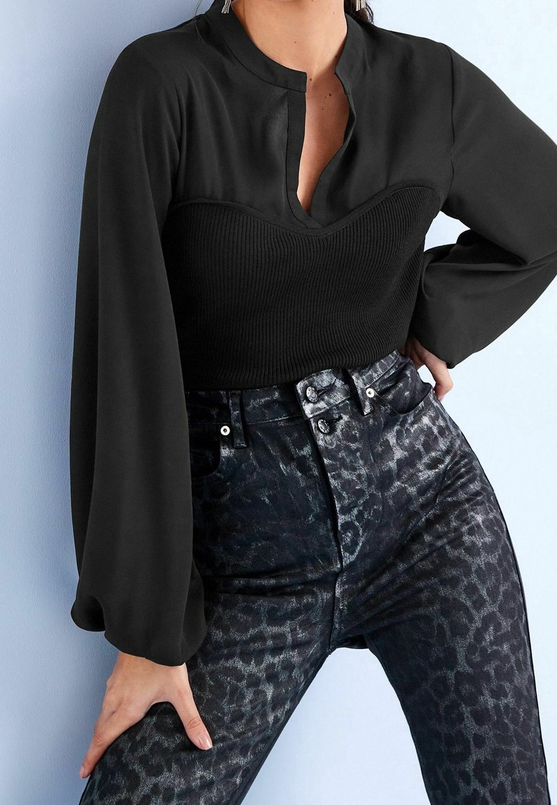 Blouse noire avec un col haut et un devant côtelé, associée à un jean noir ajusté arborant un imprimé léopard discret et une texture brillante.