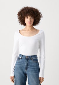 Blusa blanca de manga larga con escote amplio, combinada con unos vaqueros azules de cintura alta con detalles en tono dorado y un corte clásico.