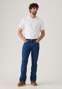Vaqueros de mezclilla azul con un corte recto, combinados con una camiseta blanca de cuello redondo. El hombre lleva zapatos de cuero marrón y las manos en los bolsillos.