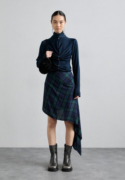 Top blu navy a maniche lunghe arricciate con dettagli a bottoni, abbinato a una gonna asimmetrica a quadri verde e blu navy e stivaletti alla caviglia grigi. Borsa rotonda nera.
