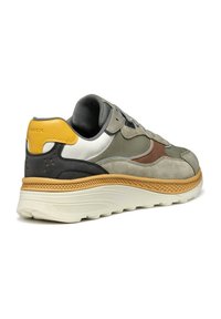 Sneaker grigio chiaro realizzato in suede e mesh, con un design nero e marrone, un accento giallo sul tallone e una suola bianca texture.