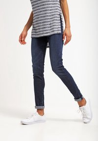 Mörkblå skinny jeans med upprullade cuffs, kombinerade med vita sneakers. Tyget har en slät yta och subtila sömnadsdetaljer.