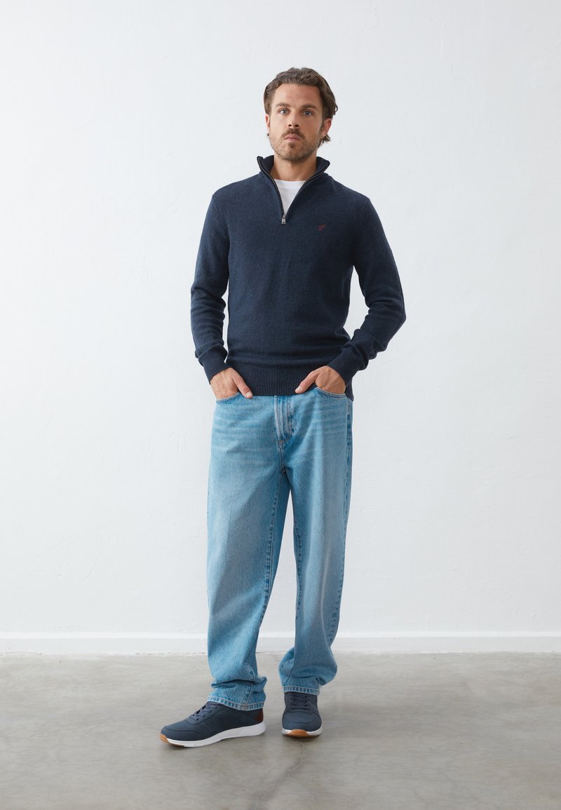 Navy zip-up sweater with ribbed cuffs and hem, paired with light blue jeans and dark sneakers. Minimal branding on the sweater.