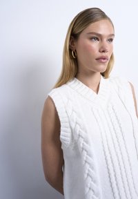 Femme blonde aux yeux bleus portant un gilet sans manches en tricot torsadé blanc et des boucles d'oreilles créoles en or, regardant vers la droite.