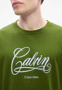 Young man wearing green Calvin Klein t-shirt with large white script text and smaller printed brand name below.
