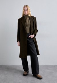 Lang donker groen wollen jas met oversized revers, gecombineerd met een aansluitende olijfgroene coltrui en wijde zwarte jeans, en bruine chunky laarzen.