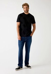 Zwart T-shirt met een subtiel patroon, gecombineerd met een blauwe spijkerbroek. Het model draagt zwart-witte sneakers. Simpele casual look.