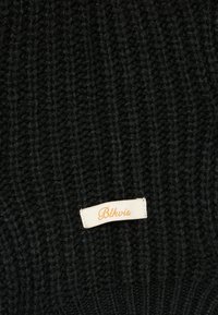 Μαύρος ριγέ πλεκτός ύφασμα με μια μικρή λευκή ορθογώνια ετικέτα ραμμένη, που φέρει πορτοκαλί καλλιγραφικό κείμενο που λέει "Blkvis".