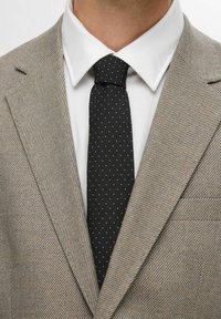 Beige fiskebensmønstret dress med skrå revers, parret med en hvid skjorte og et sort slips med hvide prikker, vist ved overkroppen.