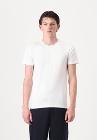 Balta medvilninė marškinėliai su apvalia iškirpte ir trumpomis rankovėmis, pasižymintys lygiu tekstūru ir prigludusiu dizainu, dėvimi su tamsiomis kelnėmis.