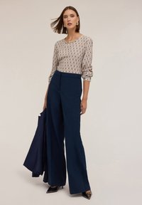 Pantaloni a gamba larga di colore blu navy abbinati a una blusa crema con motivi geometrici e un collo decorativo, completati da tacchi neri.