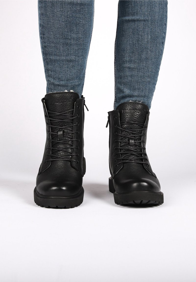 Personne portant des bottes noires en cuir à lacets et un jean bleu en denim, debout sur une surface blanche.