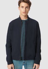 Giubbotto bomber navy con colletto alto, zip frontale e tasche laterali, indossato sopra una camicia blu strutturata, abbinato a jeans chiari.
