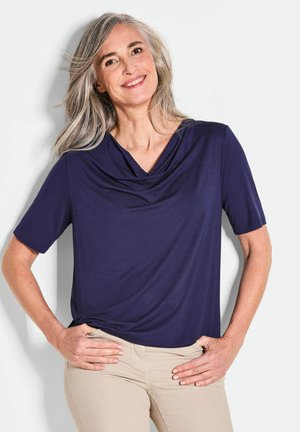 Top blu navy con maniche corte, realizzato in tessuto morbido. Presenta un colletto drappeggiato e una vestibilità comoda, abbinato a pantaloni beige chiaro.
