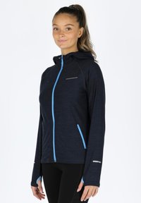 Giacca con cappuccio blu navy con accenti azzurri, dotata di chiusura con zip e tasche laterali. Realizzata in un tessuto strutturato e elasticizzato.