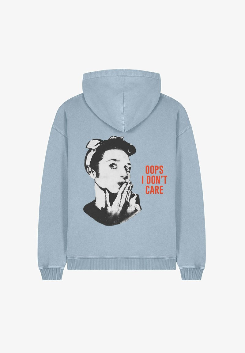 Hoodie bleu clair avec un graphisme noir et blanc représentant une femme faisant un geste de silence et un texte rouge : "OUPS, JE M'EN FOUS." Tissu doux.
