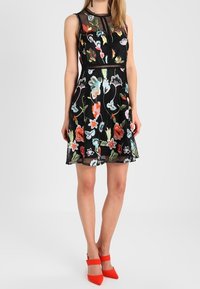 Czarna sukienka bez rękawów z kolorowym haftem kwiatowym, dopasowana górna część i rozkloszowany dół. Czerwone szpilki w szpic dopełniają stylizację.