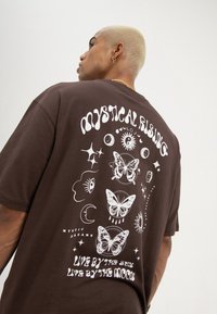 Person with short blonde hair wearing a brown shirt with white mystical designs, butterflies, and text reading "Mystical Rising" and "Live by the sun."