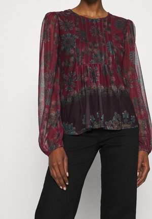 Długi rękaw, przezroczysta bordowa bluzka w kwiaty z plisami i marszczonym pasem, noszona z czarnymi spodniami.