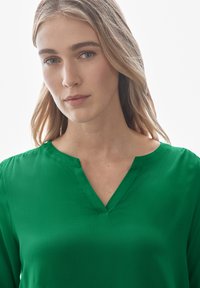 Blusa verde a maniche corte con scollo a V, realizzata in tessuto leggero e liscio, con spalle strutturate e vestibilità rilassata.