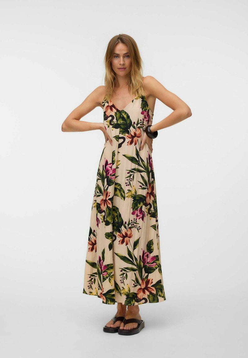 Femme debout, les mains sur les hanches, portant une robe longue beige avec des motifs floraux tropicaux colorés et des sandales à plateformes noires.