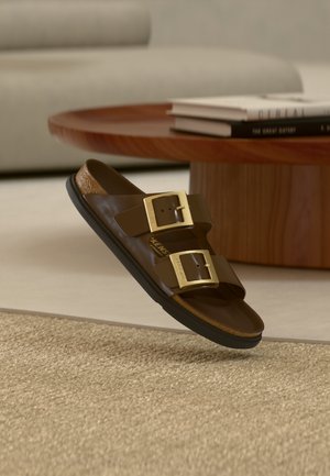 Braune Ledersandale mit zwei goldenen Schnallen, schwarzer Sohle, liegend in der Nähe eines hölzernen Couchtisches mit gestapelten Büchern in einem beigen Wohnzimmer.