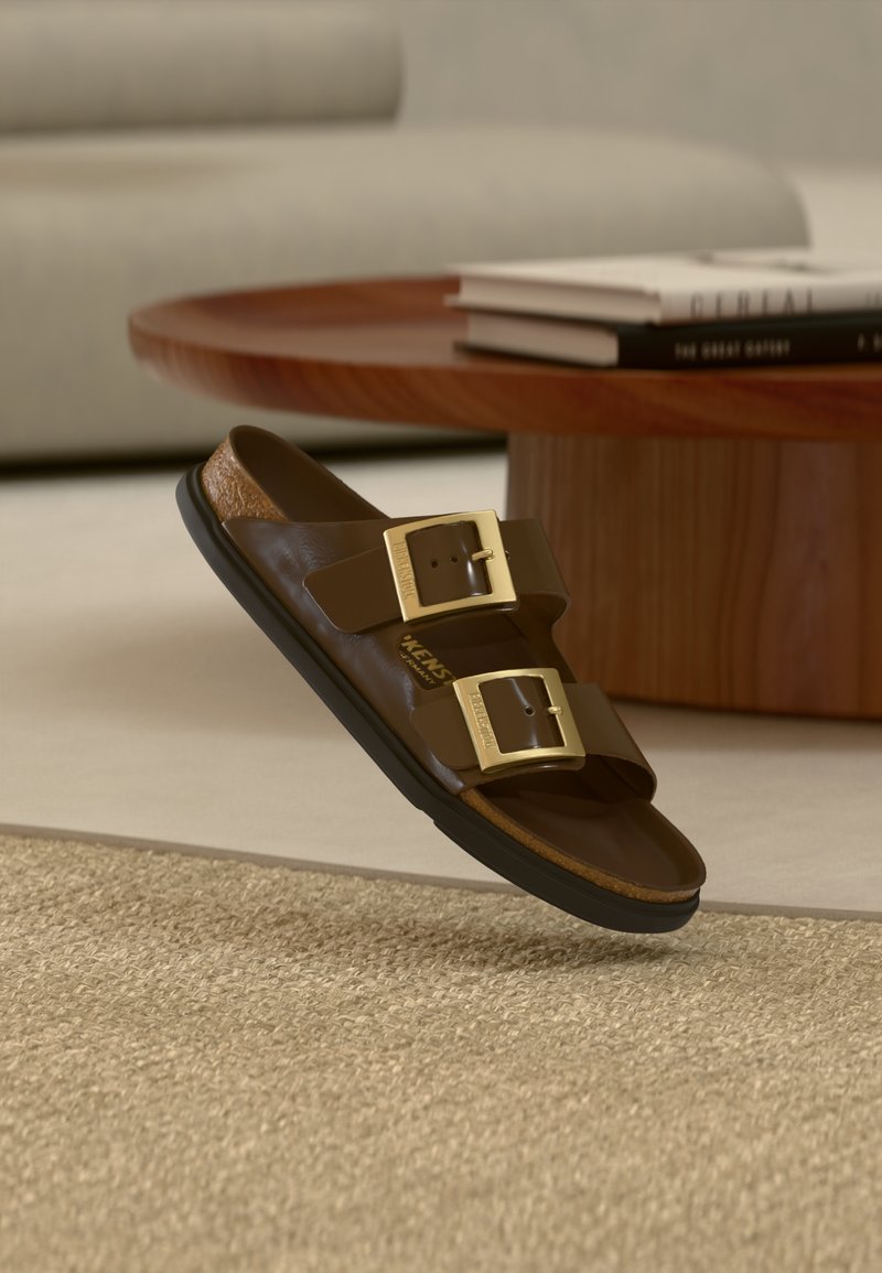 Καφέ δερμάτινο σανδάλι slide με δύο χρυσές αγκράφες, μαύρη σόλα, τοποθετημένο κοντά σε ξύλινο τραπεζάκι σαλονιού με στοιβαγμένα βιβλ ία σε μπεζ σαλόνι.