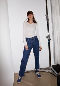 Langärmliges, weißes Oberteil kombiniert mit lockeren blauen Jeans und blauen Sneakers. Eine braune Kappe wird getragen und ein chromfarbener Hintergrund ist sichtbar.