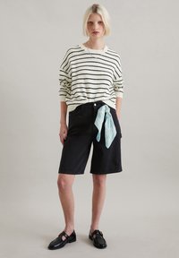 Gestreifter Strickpullover in Creme und Schwarz, kombiniert mit schwarzen Denim-Shorts und einem hellblauen, gemusterten Schal, der um die Taille gebunden ist. Schwarze Schuhe.
