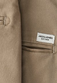 Μπεζ ύφασμα με ραμμένη τρύπα κουμπιού και μια μικρή λευκή ετικέτα που γράφει "JACK & JONES EST. 1990" δίπλα σε ένα λεπτό άνοιγμα τσέπης.