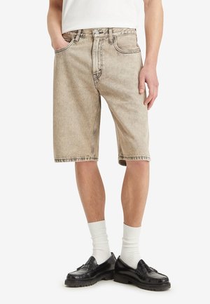 Homme portant un short en jean beige jusqu'aux genoux, des chaussettes blanches et des mocassins en cuir noir, debout avec une main dans la poche.