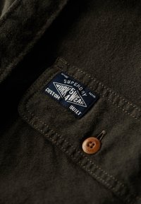 Donkergroene stof met een gestructureerd oppervlak, met een blauw-witte logo patch en een bruine knoop op de borstzak.
