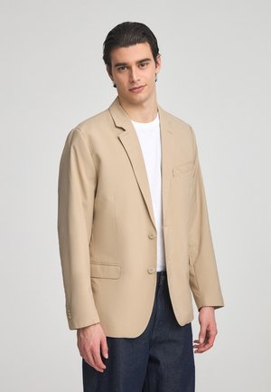 Blazer beige realizzato in un tessuto liscio, con due bottoni, revers classici e una singola tasca sul petto, abbinato a una maglietta bianca.