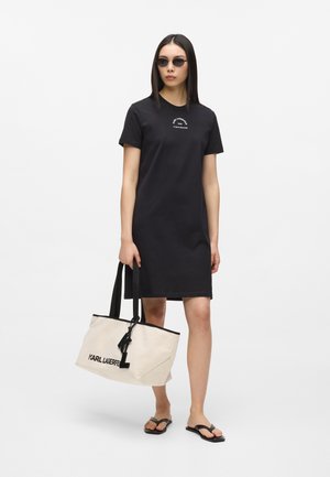 Žena u crnoj haljini s kratkim rukavima i sunčanim naočalama drži veliku bež torbu s crnim ručkama i natpisom, nosi crne japanke.