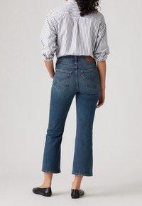 Jean en denim bleu stretch avec une taille haute, une jambe évasée et un ourlet brut. Associé à une chaussure plate noire et une chemise à rayures boutonnée.