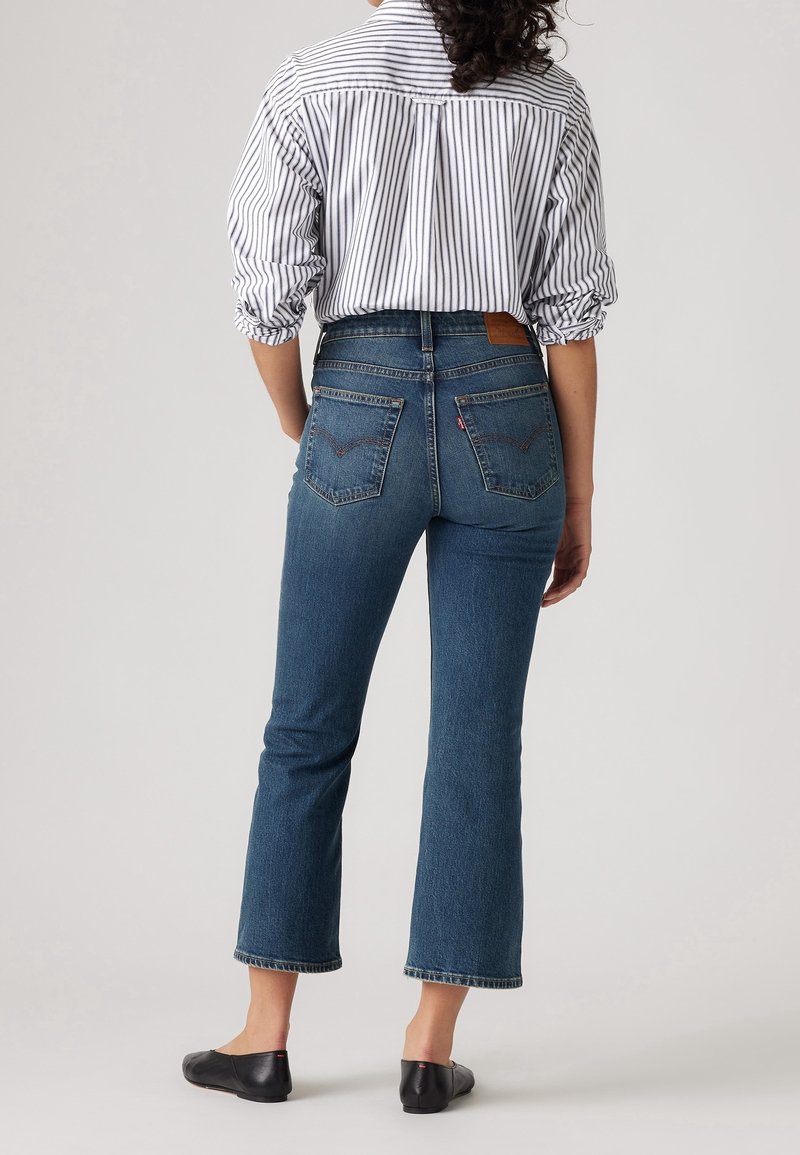 Jean en denim bleu stretch avec une taille haute, une jambe évasée et un ourlet brut. Associé à une chaussure plate noire et une chemise à rayures boutonnée.