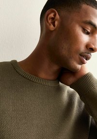 Maglione lavorato a maglia verde oliva con scollo rotondo e polsini a coste, che mostra un tessuto strutturato e una vista ravvicinata del colletto e della spalla.