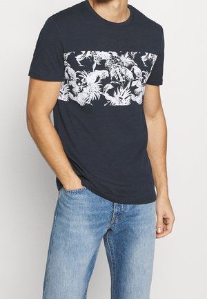 Hombre con camiseta azul marino oscuro con banda blanca con estampado tropical en el pecho y vaqueros azul claro, de pie con una mano en el bolsillo.