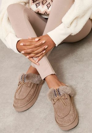 Pantofi slip-on din suede bej, cu șnururi din blană artificială, cusături decorative și accente împletite contrastante la deschideri. Textură moale și talpă căptușită.