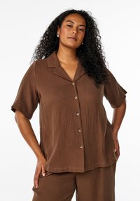 Woman with long curly hair wearing brown short-sleeve button-up shirt and matching pants, posing with hands on hips against white background.