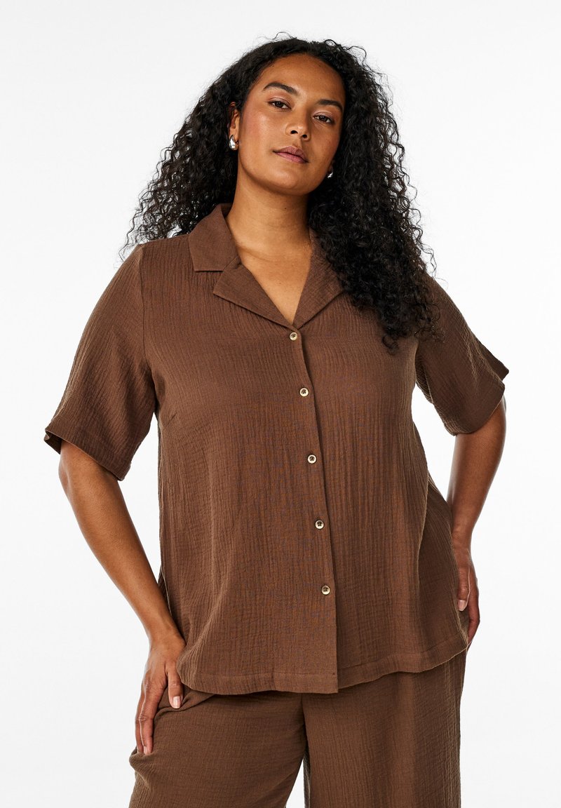 Woman with long curly hair wearing brown short-sleeve button-up shirt and matching pants, posing with hands on hips against white background.