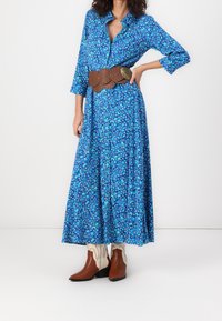 Langes blaues Blumenkleid mit Knopfverschluss, Dreiviertelärmel, getragen mit einem braunen Gürtel mit kreisförmigen Ausschnitten und braunen Stiefeln.